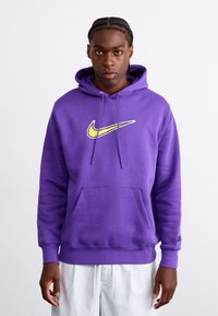 Violetinis džemperis su gobtuvu, pagamintas iš minkštos medvilnės, su geltonu Nike logotipu ant krūtinės, priekinė kišenė ir raišteliai ant gobtuvo.