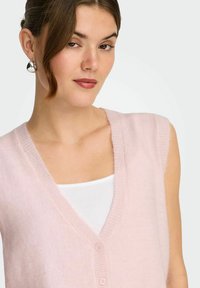 Jeune femme aux cheveux bruns portant des boucles d'oreilles argentées, un haut blanc et un gilet sans manches rose pâle à boutons, sur un fond uni.