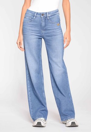 Persona che indossa jeans ampi color azzurro chiaro e scarpe da ginnastica bianche, in piedi contro uno sfondo chiaro e uniforme.