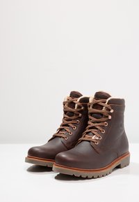 Botas de tornozelo em pele castanha com forro de pele sintética bege, cordões castanhos e solas de borracha robustas. Apresenta ilhós de metal e um design cosido.