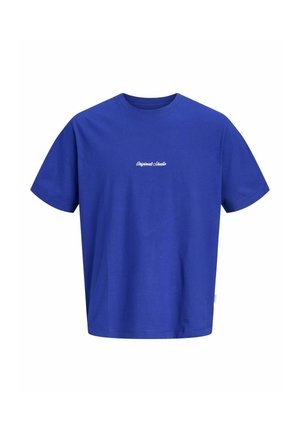 Młoda osoba stojąca, ubrana w fioletowy t-shirt z napisem, dżinsowe szorty, białe skarpetki i białe trampki na jednolitym tle.