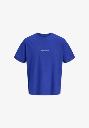 Młoda osoba stojąca, ubrana w fioletowy t-shirt z napisem, dżinsowe szorty, białe skarpetki i białe trampki na jednolitym tle.