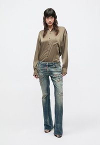 Svetlohnedá saténová košeľa na gombíky v kombinácii s modrými zvonovými džínami, zdobená ozdobnými kameňmi a vyblednutím. Model stojí na obyčajnom pozadí.