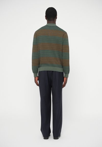 Zaļš un brūns svītrots adīts kardigans ar ribotu aproci un apakšmalu, kombinēts ar tumši zilām svītrainām biksēm; skatīts no mugurpuses.