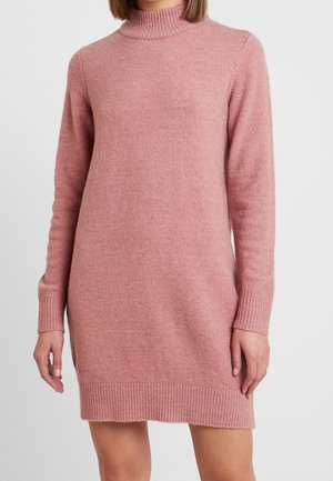 Kobieta w długim rękawie, różowej sukience sweterkowej o wysokim kołnierzu, z prążkowanymi mankietami i brzegiem.