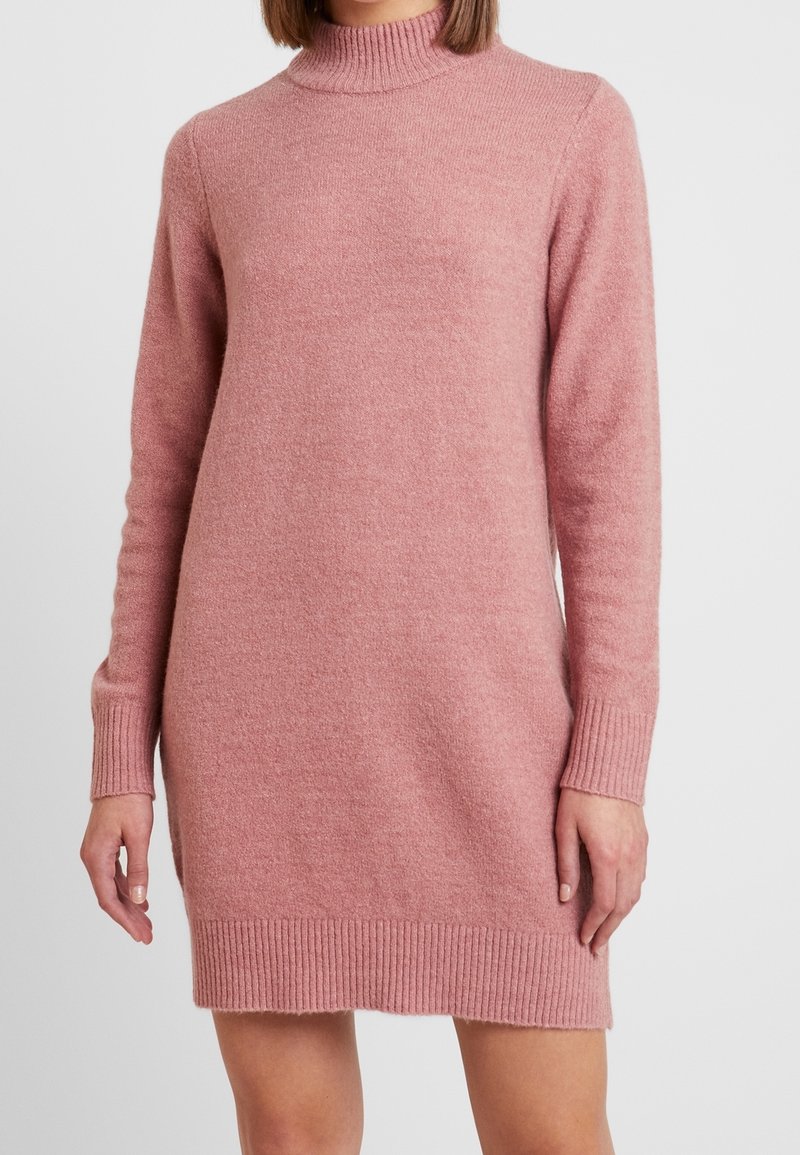 Femme portant une robe en tricot rose poussiéreux à manches longues, col haut, avec des poignets et un ourlet côtelés.