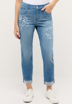 Blaue, aufgerollte Jeans mit weißen Grafiken von Cupido und Herzpfeil, Schriftzug "be mine", getragen mit weißen Turnschuhen und einem eingesteckten Oberteil.