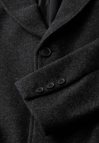 Vlněný kabát tmavě šedé barvy s klopami, vybavený hladkou podšívkou a čtyřmi černými knoflíky na manžetách. Viditelná strukturovaná tkanina.