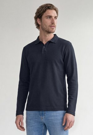 Homme aux cheveux courts et ondulés portant un polo à manches longues foncé et un jean bleu clair, debout et regardant vers la droite.