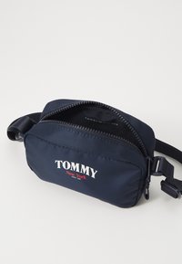 Marineblå nylon crossover-veske med glidelås, med frontlogoen "TOMMY New York" i hvitt og rødt, flat form med justerbar stropp.