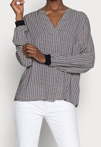 Blouse à manches longues en motif géométrique noir et blanc, avec un décolleté en V et des poignets noirs, fabriquée en tissu lisse. Portée avec un pantalon blanc.