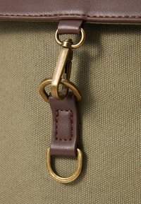 Dettaglio di una borsa in tela con una chiusura in bronzo, accenti in pelle marrone e materiale oliva texturizzato. L'hardware notevole include un grande anello metallico.