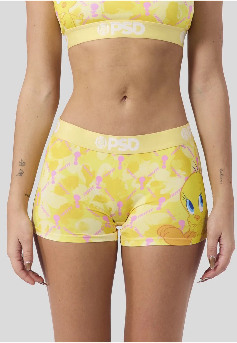 Femme portant un crop top jaune et un short avec un motif de Titi, debout les bras détendus de chaque côté sur un fond gris clair.