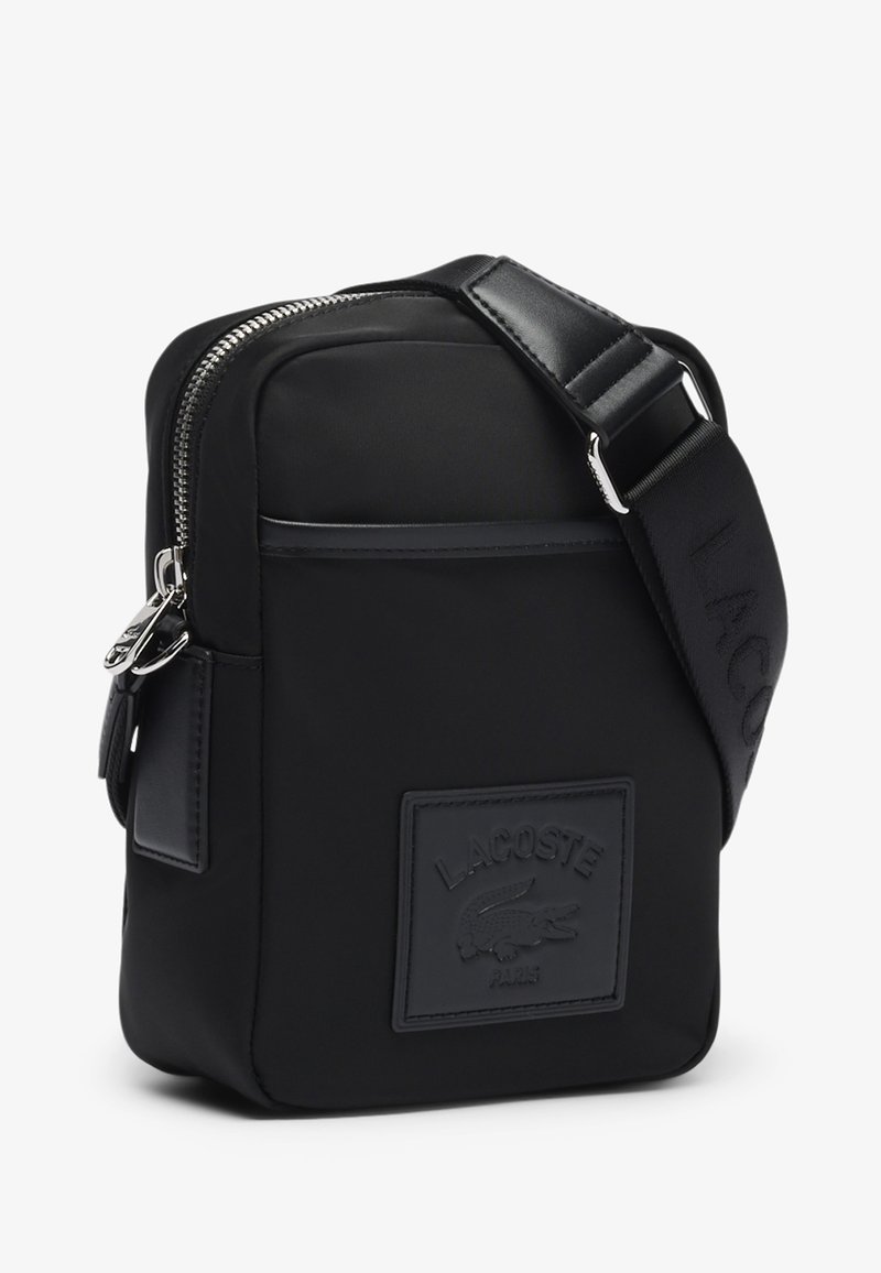 Sac bandoulière noir en matériau synthétique. Dispose d'une fermeture éclair, d'une sangle réglable et d'un patch logo texturé à l'avant. Design compact.