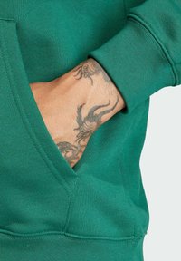Zelená mikina s predným vreckom, rebrovanými manžetami a mäkkou textúrou. Viditeľné tetovanie na ruke s komplikovaným dizajnom.