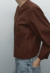 Brunstribet skjorte med rundet krave, knaplukning foran og lange ærmer. Stoffet ser glat ud og er parret med lyseblå jeans.