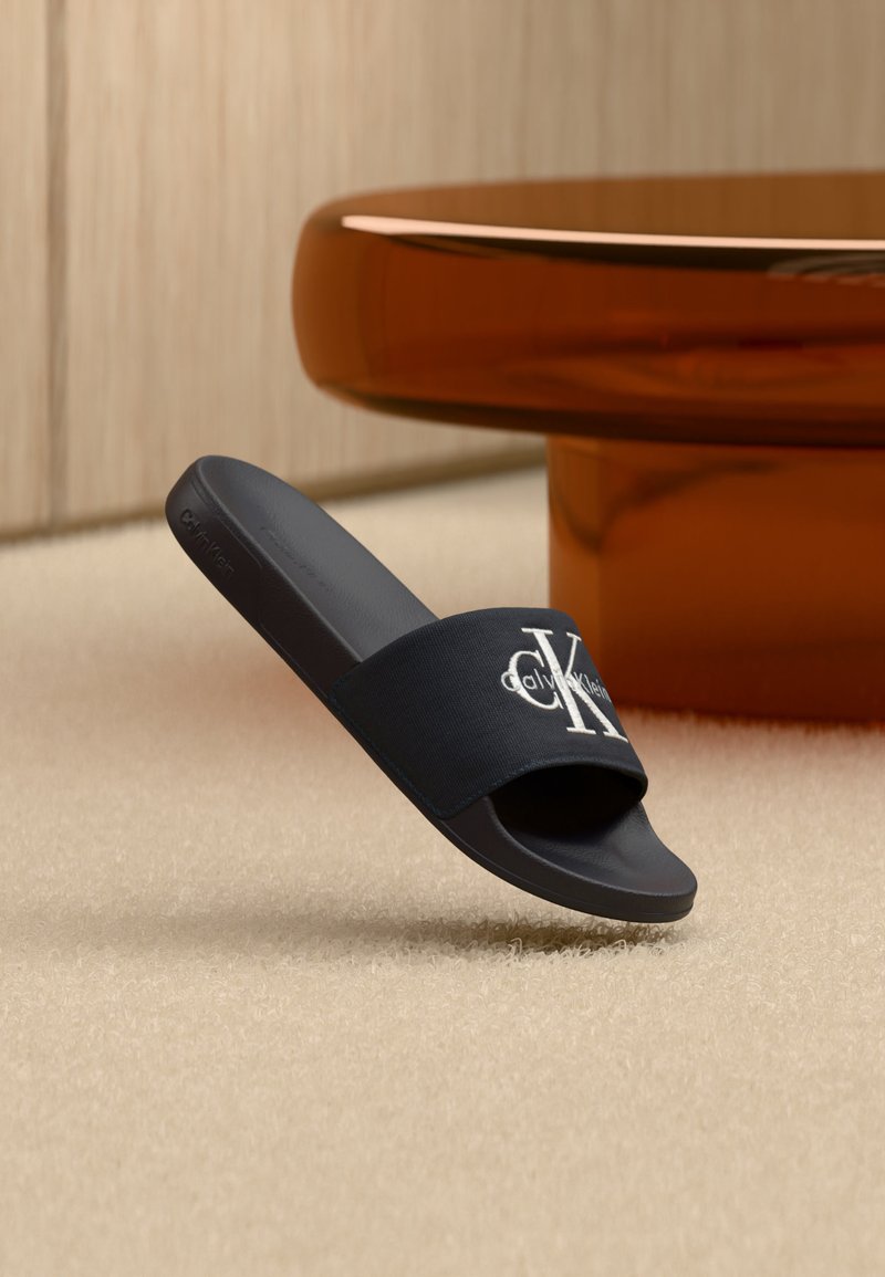 Μαύρο σανδάλι Calvin Klein τύπου slide με λευκό λογότυπο στο λουρί, αιωρούμενο πάνω από μπεζ χαλί κοντά σε στρογγυλό, διάφανο καφέ τραπέζι.