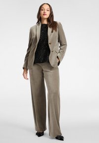 Completo in velluto grigio-taupe tenue, composto da blazer monopetto con bottoni dorati e pantaloni a gamba larga, abbinato a una maglia nera strutturata.