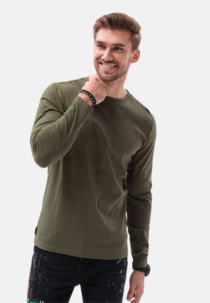 Camisa de manga larga color verde oliva hecha de algodón, con un escote redondo. Combinada con unos jeans oscuros que muestran salpicaduras multicolores.