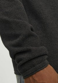 Maglione scuro e testurizzato con un motivo lavorato a maglia. I polsini sono arrotolati, evidenziando una vestibilità aderente e un orlo finito.