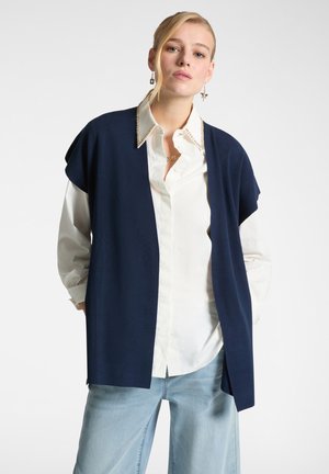 Cardigan senza maniche blu navy indossato sopra una camicia bianca con colletto decorativo; abbinato a jeans a gamba larga azzurri.