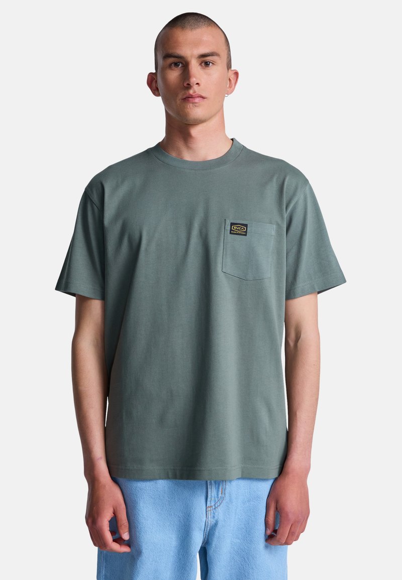 Joven con cabello corto, vistiendo una camiseta verde de cuello redondo con bolsillo en el pecho y jeans azul claro, de pie frente a un fondo blanco.