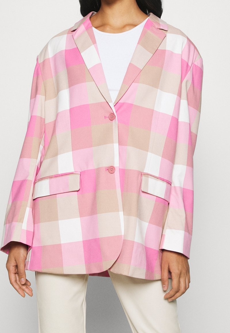 Femme portant un blazer oversize à carreaux rose, beige et blanc avec deux boutons et des poches à rabat sur une chemise blanche.