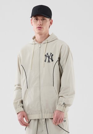 Joven con sudadera con capucha beige y pantalones con ribete negro y una gorra de béisbol negra con el logo de los New York Yankees.