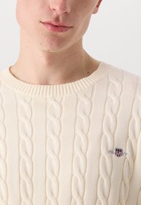 Light beige cable-knit sweater featuring a ribbed neckline and a small embroidered logo on the left chest.