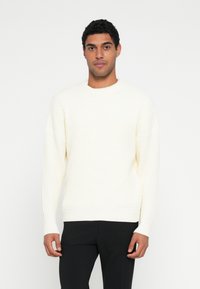 Kermanvärinen neulepusero, jossa teksturoitu kuvio, pyöreä pääntie ja resorit hihansuissa. Yhdistetty mustiin housuihin.