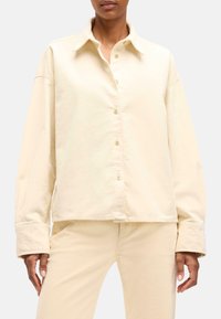 Camicia button-up di un beige chiaro, con una texture morbida, design con colletto e maniche lunghe con polsini larghi. Abbinata a pantaloni coordinati di un beige chiaro.