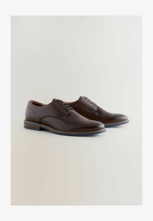 Tumši brūnas ādas kurpes ar gludu virsmu, šņorēm un apaļu purngalu. Kurpēm ir šūta zole un neliels papēdis atbalstam.