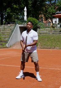 White polo shirt with gray accents, gray shorts, and white sneakers with black stripes, holding a tennis racket on a clay court.