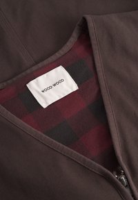 Rjav zip-up kos, ki ima podlogo v karirastem vzorcu burgundske in črne barve. Etiketa glasi "WOOD WOOD." Tkanina ima teksturiran videz.