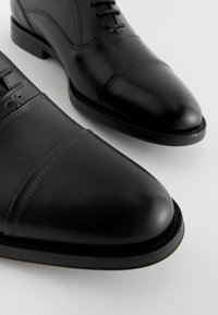 Schwarze Lederschuhe mit glatter Oberfläche, mit Kappen-Design und subtilen Steppnähten, runden Zehen und flachen Sohlen.