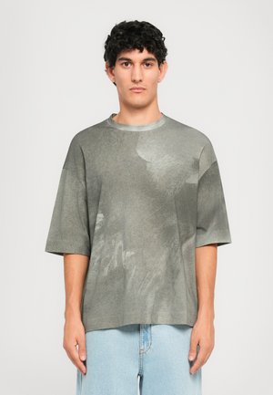 Junger Mann, der ein übergroßes, graues Batik-T-Shirt mit kurzen Ärmeln und hellblauen Jeans trägt, im Stehen vor einem schlichten weißen Hintergrund.