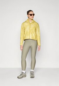 Geltona, dažyta kaklaraiščiu striukė su aukštu apykakliu ir užtrauktuku, derinama su šviesiai alyvuogių spalvos tamprėmis ir pilkais sportiniais bateliais. Dėvimos saulės akiniai.