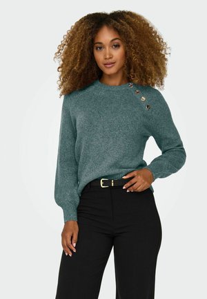 Femme aux cheveux bouclés portant un pull en maille vert avec des boutons aux épaules et un pantalon noir avec ceinture noire, debout devant un fond uni.