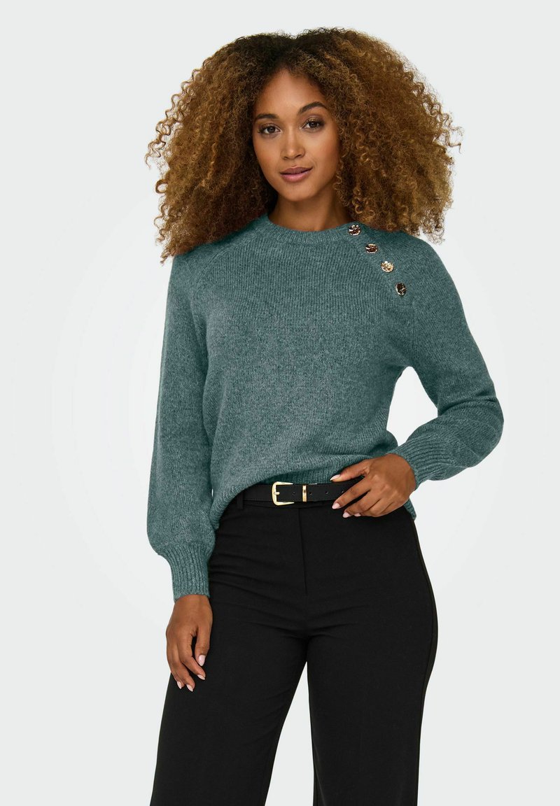 Femme aux cheveux bouclés portant un pull en maille vert avec des boutons aux épaules et un pantalon noir avec ceinture noire, debout devant un fond uni.