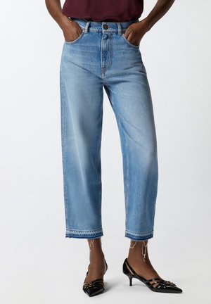 Persona che indossa jeans azzurri corti con orli grezzi e décolleté neri a punta con tacco kitten, mani in tasca su sfondo neutro.