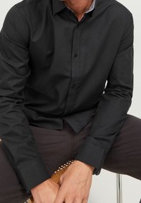 Svart långärmad skjorta med en strukturerad krage, med en subtil textur och knappstängning, kombinerad med mörka mönstrade byxor.