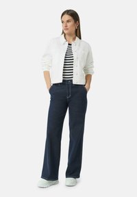 Witte denimjacket over een zwart-wit gestreepte shirt, gecombineerd met donkerblauwe wijde jeans en lichtkleurige sneakers.