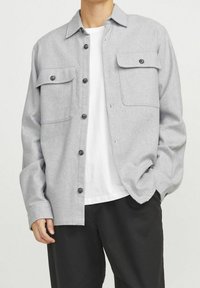 Šedá flanelová košile s knoflíkovým zapínáním, dvěma předními kapsami a klasickým límečkem, ve spojení s bílým tričkem a černými kalhotami.