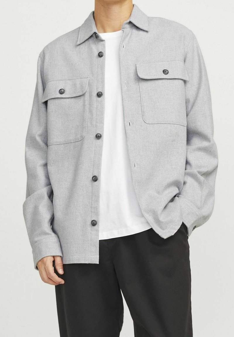 Šedá flanelová košile s knoflíkovým zapínáním, dvěma předními kapsami a klasickým límečkem, ve spojení s bílým tričkem a černými kalhotami.