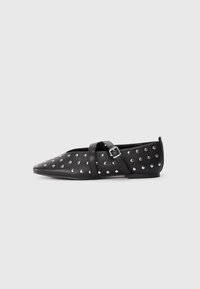 Scarpa piatta nera a punta con borchie argentate e cinturino incrociato regolabile con fibbia, tacco minimal, su sfondo chiaro uniforme.