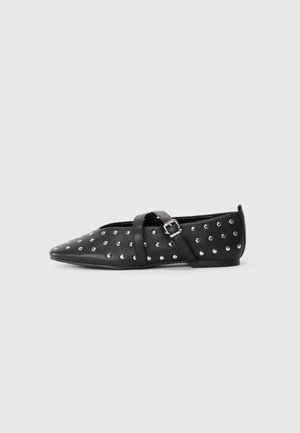 Scarpa piatta nera a punta con borchie argentate e cinturino incrociato regolabile con fibbia, tacco minimal, su sfondo chiaro uniforme.