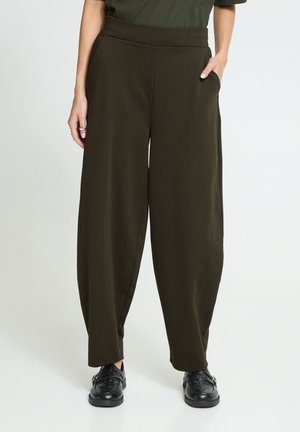 Femme portant un pantalon large olive foncé avec poches latérales et chaussures en cuir noir, debout devant un fond neutre.