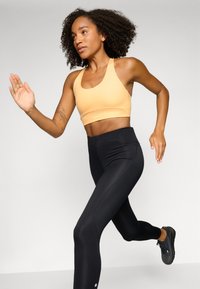 Conjunto deportivo que incluye un sujetador deportivo de color naranja claro y leggings negros de cintura alta. Los leggings tienen una textura suave y un ligero brillo.