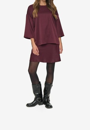 Kvinde iført en løs, bordeaux top med lange ærmer og matchende nederdel, sorte strømpebukser og sorte læderstøvler til midt på læggen med spænder.