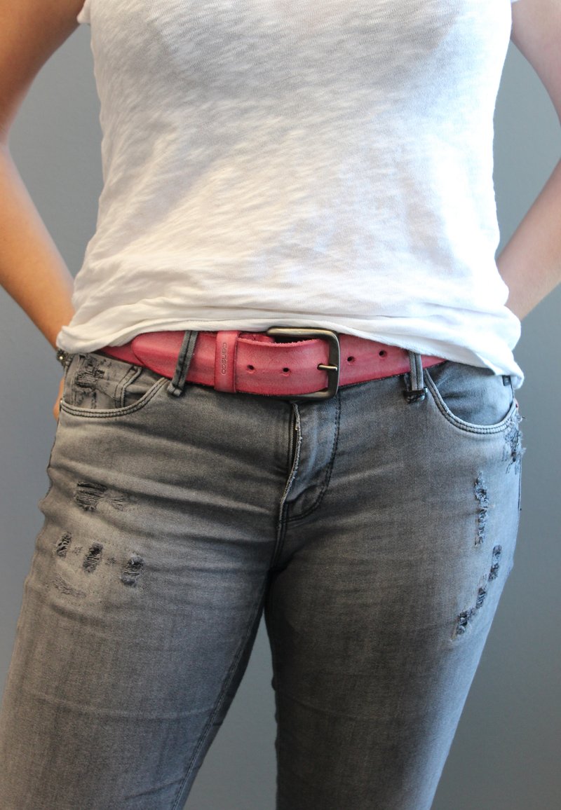 Rosa Ledergepäck mit einer metallischen Schnalle, getragen über lässigen grauen Jeans. Die Jeans weisen Risse und eine ausgewaschene Textur auf.